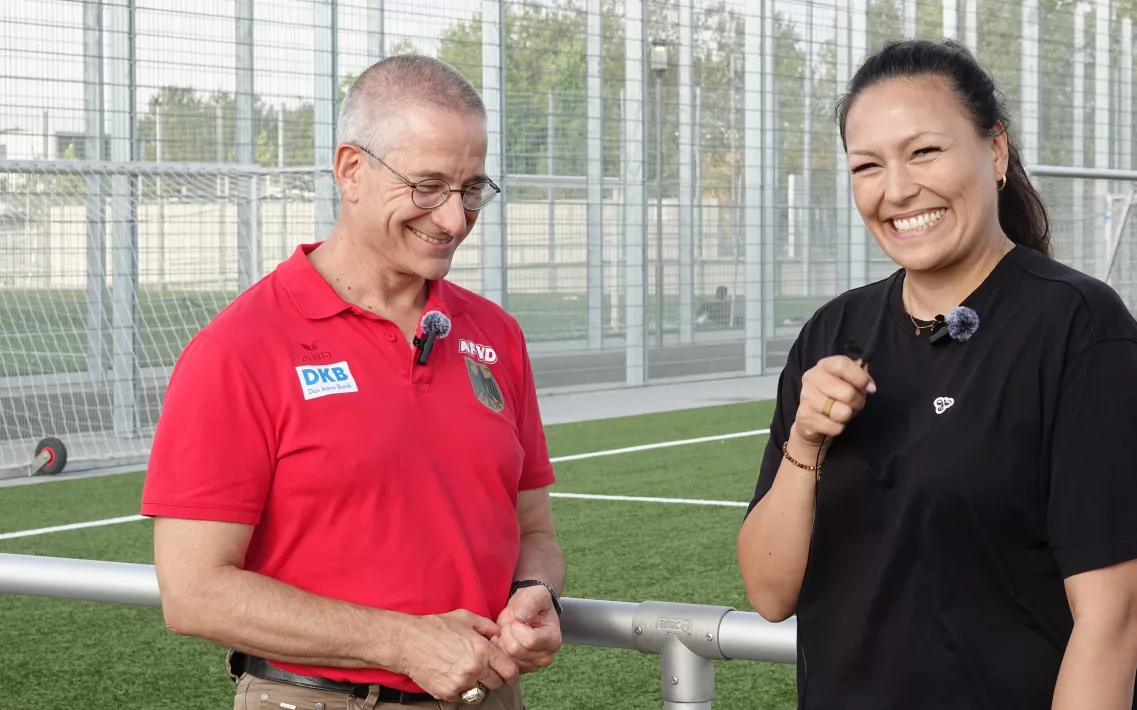 Florian Berrenberg und Nadine Nurasyid lächeln während eines Interview-Gespräches, vor einem Kunstrasensportplatz